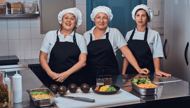 equipe-de-cozinha-feliz-vestindo-um-uniforme-ao-lado-de-uma-mesa-com-legumes-picados-posando-para-uma-camera-na-cozinha-de-um-restaurante